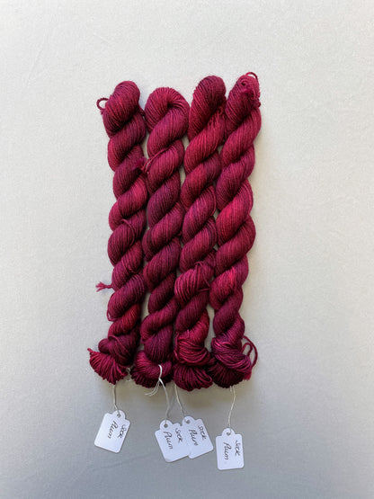 Full shot of 20g Sock weight mini skeins in semi-solid colourway Plum an intense, deep Plum/Burgundy. Photographed on a light grey background.