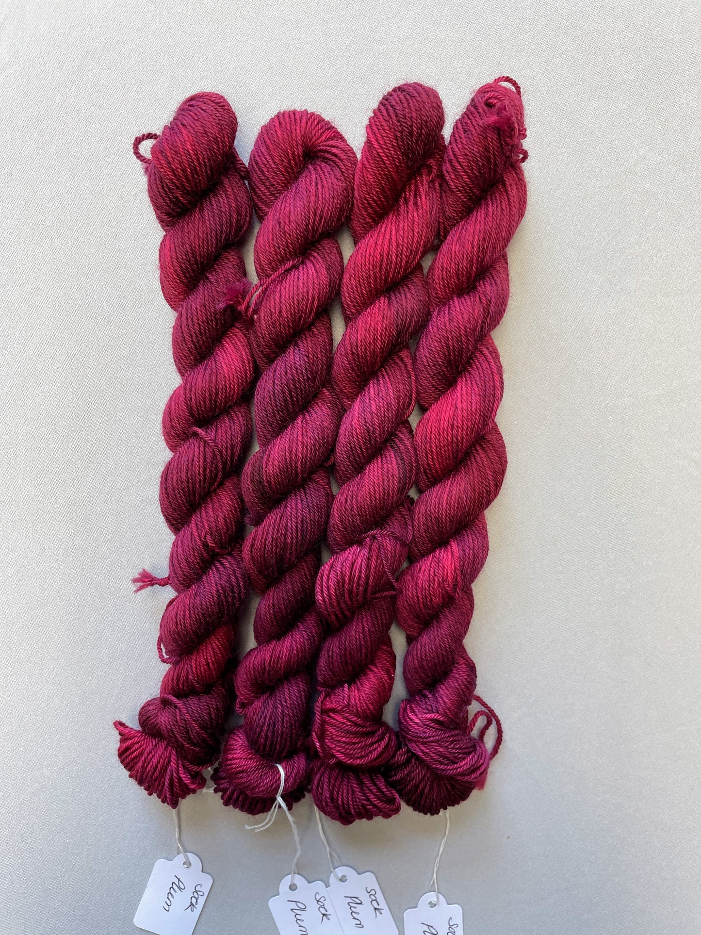 Zoomed in shot of 20g Sock weight mini skeins in semi-solid colourway Plum an intense, deep Plum/Burgundy. Photographed on a light grey background.