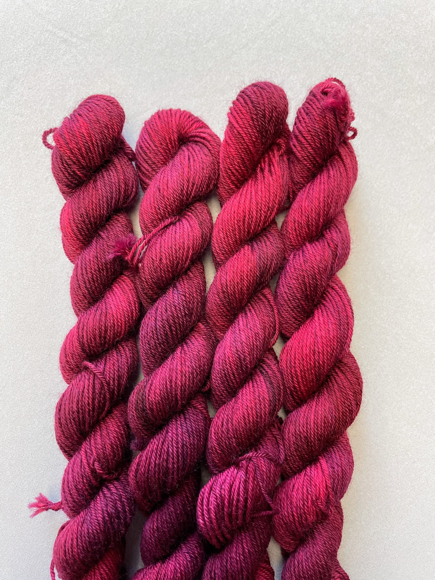 Close up shot of 20g Sock weight mini skeins in semi-solid colourway Plum an intense, deep Plum/Burgundy. Photographed on a light grey background.