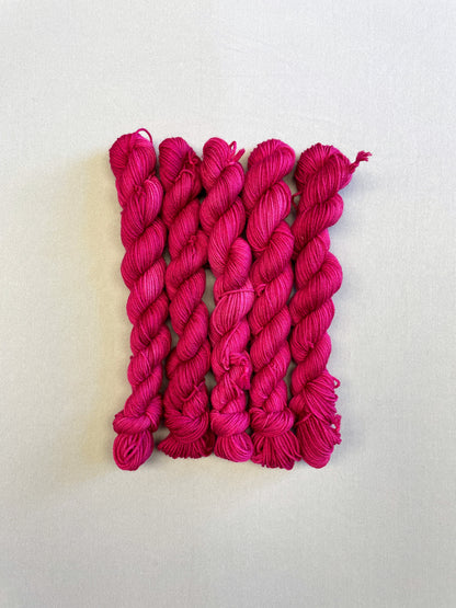 Full shot of 20g sock weight mini skeins in semi-solid colourway Raspberry, an intense, deep pink. Photographed on a light grey background.