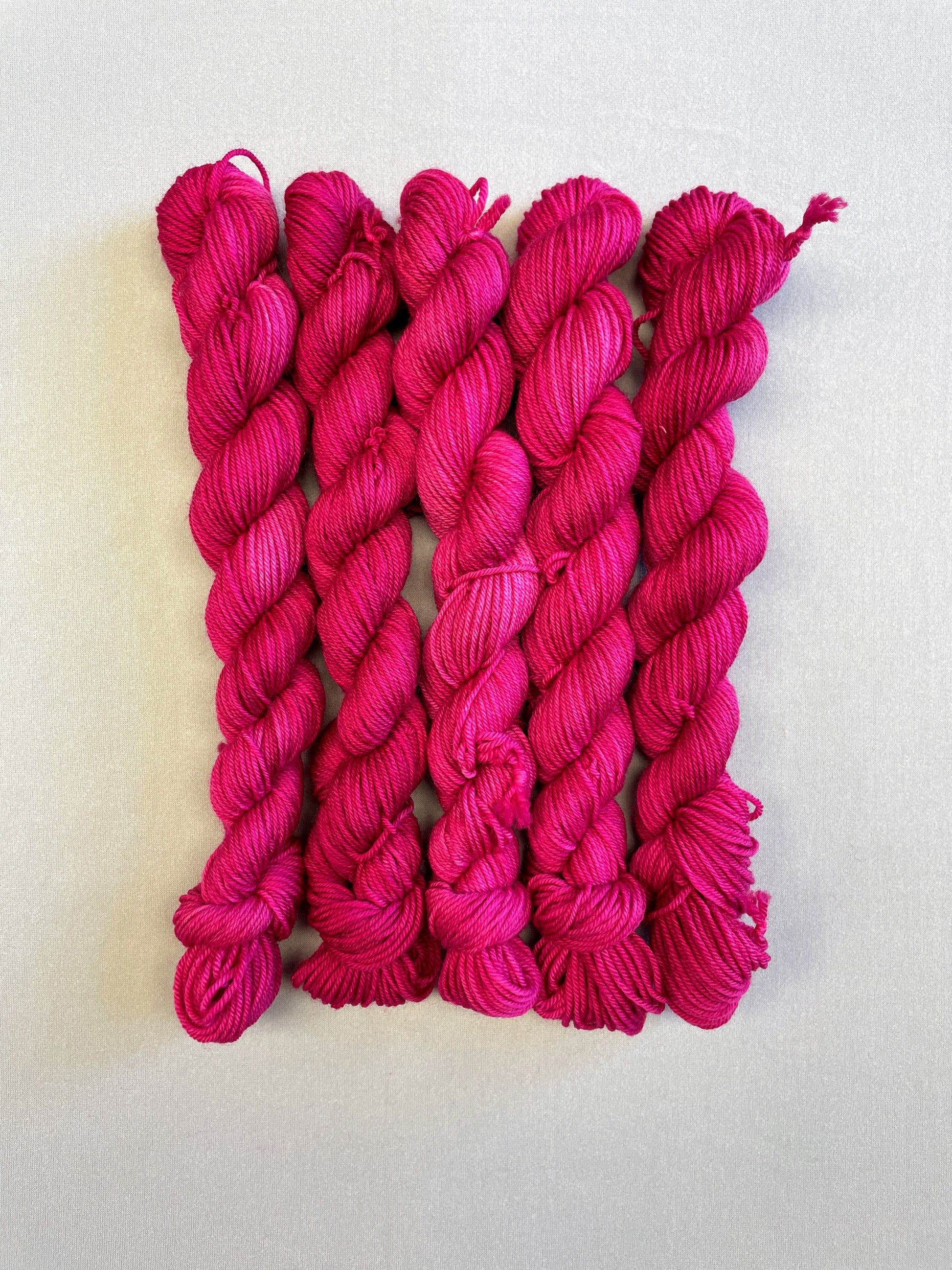 Zoomed in shot of 20g sock weight mini skeins in semi-solid colourway Raspberry, an intense, deep pink. Photographed on a light grey background.