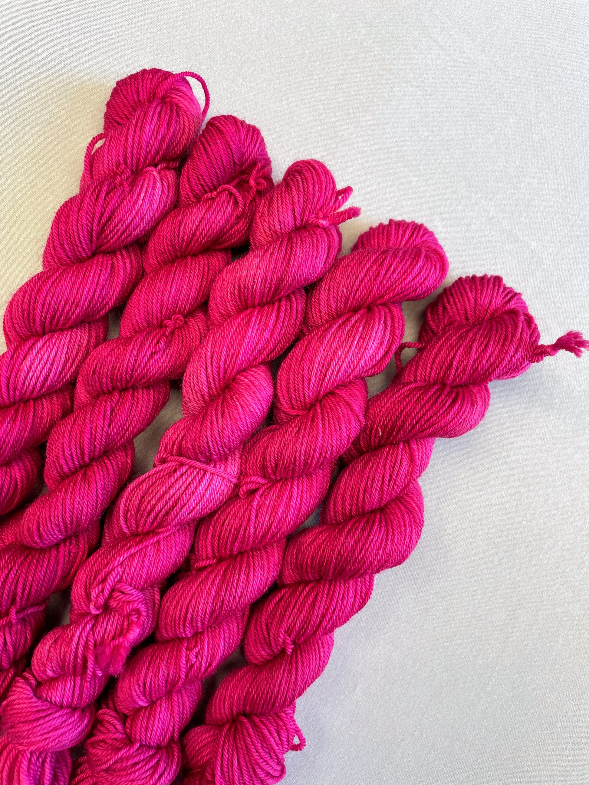 Close up shot of 20g sock weight mini skeins in semi-solid colourway Raspberry, an intense, deep pink. Photographed on a light grey background.