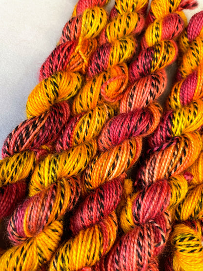 Hand-dyed Yarn
Close up shot of 5 x 20g twisted, Zebra Sock mini skeins in colourway Phoenix, photographed on light grey background. This colourway is full of tones of Golden Poppy, Ruby Red and Saffron with the black and white strip running through it. 