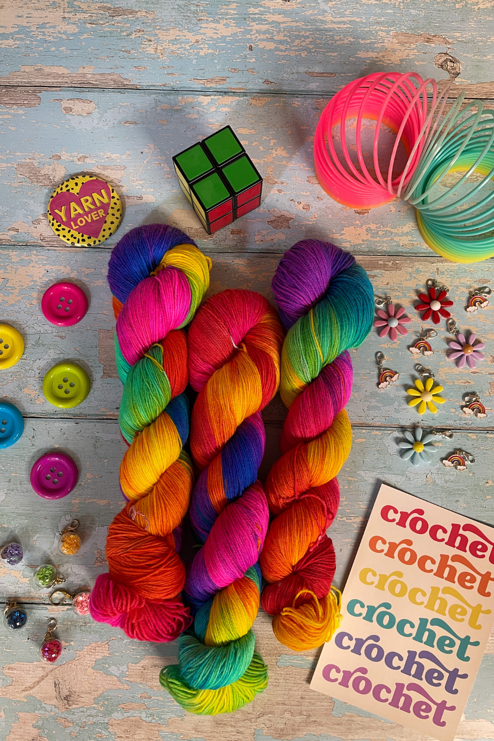 3 twisted skeins of Hand-dyed Yarn in colourway Bright Rainbow. The yarn contains 7 bright colours, sunshine, lava, ruby, apple, cyan, electric purple and neon pink. Surrounded by props, such as stitch markers and buttons on a blue, wooden background. 