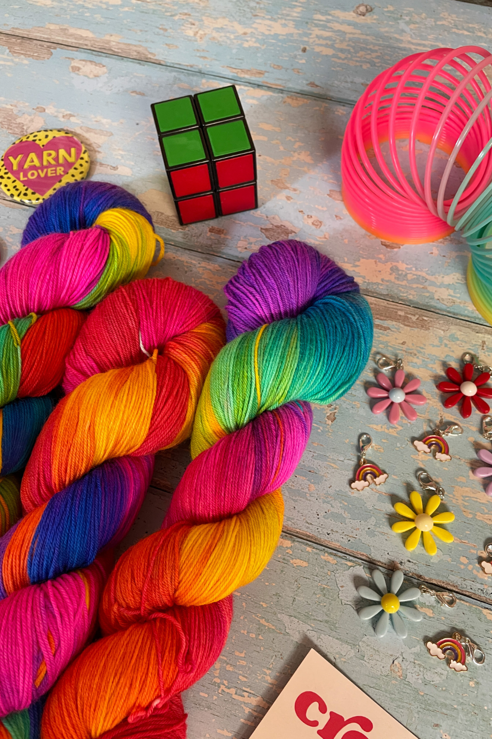 Close up photo of 3 twisted skeins of Hand-dyed Yarn in colourway Bright Rainbow. The yarn contains 7 bright colours, sunshine, lava, ruby, apple, cyan, electric purple and neon pink. Surrounded by props, such as stitch markers and rubix cube on a blue, wooden background. 