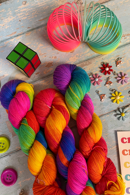 Close up photo of 3 twisted skeins of Hand-dyed Yarn in colourway Bright Rainbow. The yarn contains 7 bright colours, sunshine, lava, ruby, apple, cyan, electric purple and neon pink. Surrounded by props, such as stitch markers and slinky on a blue, wooden background. 