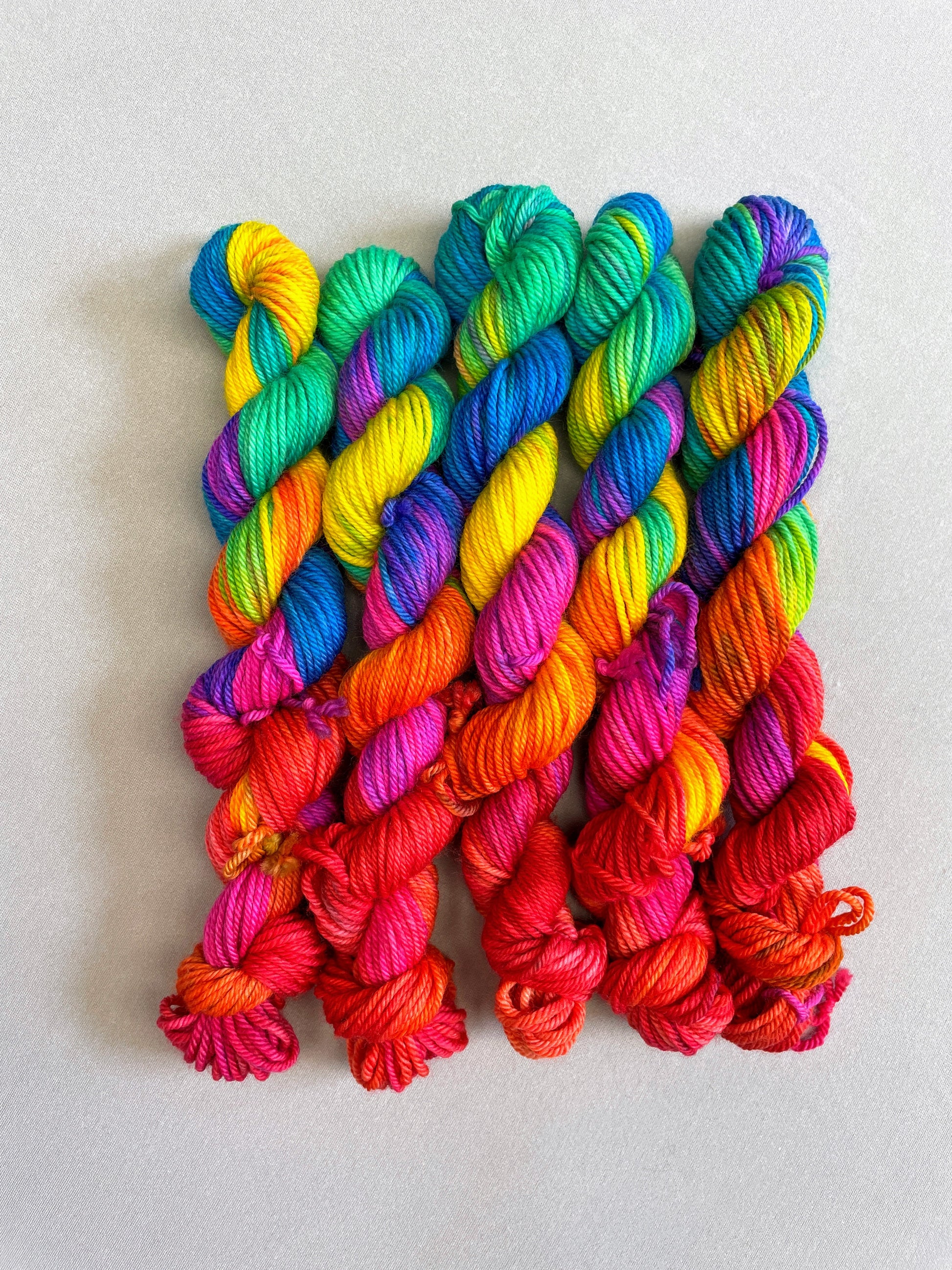 5 x 20g Hand-dyed Yarn mini skeins in colourway Bright Rainbow. The image shows the full skeins, twisted up and is on our DK weight Yarn Base. The yarn contains 7 bright colours, sunshine, lava, ruby, apple, cyan, electric purple and neon pink and is photographed on a white background.